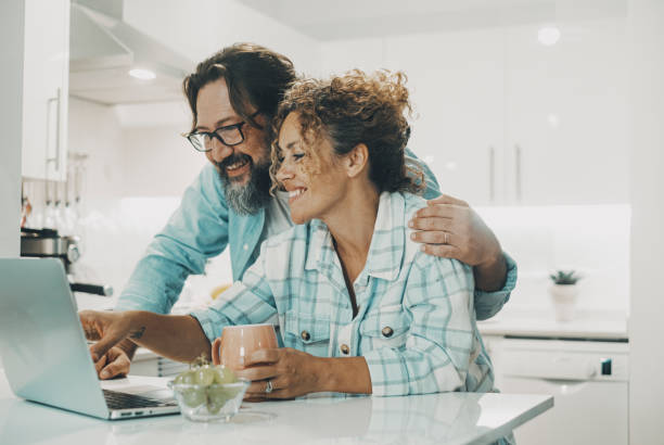 Happy husband and wife enjoying time in the kitchen using laptop computer, young couple looking at notebook screen browsing internet choosing shopping online together in apartment, People technology