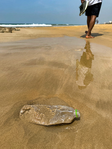 Stock photo showing close-up view of golden sandy Sri Lankan tropical beach at low tide with crushed, transparent plastic water bottle without lid containing sand and water washed up from polluted sea. Coastline view from Mount Lavinia Beach, Colombo, Sri Lanka.