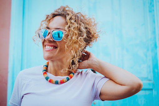 Colorful portrait of a young adult woman smiling against a blue wall background and wearing sunglasses. Positive female people. Attractive blonde mature lady enjoy and have fun alone in leisure