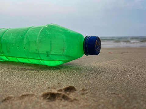 Stock photo of showing close-up view of sandy beach at low tide with empty, green plastic drinks bottle with blue lid washed up from polluted sea.