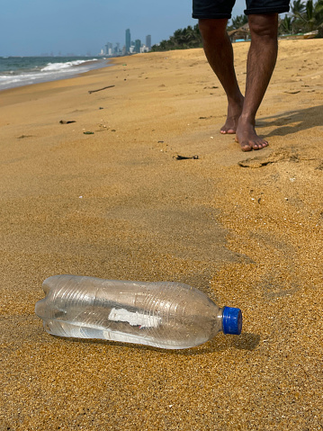 Stock photo showing close-up view of golden sandy Sri Lankan tropical beach at low tide with empty, transparent plastic drinks bottle with blue lid washed up from polluted sea. Coastline view with exotic tall coconut palm trees and a background of the city of Colombo from Mount Lavinia Beach, Colombo, Sri Lanka.
