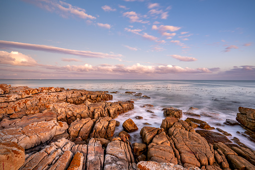 Crashing waves on the rocky shoreline of Fish Hoek with a vibrant sunset over False Bay on the Cape Peninsula outside Cape Town, South Africa.