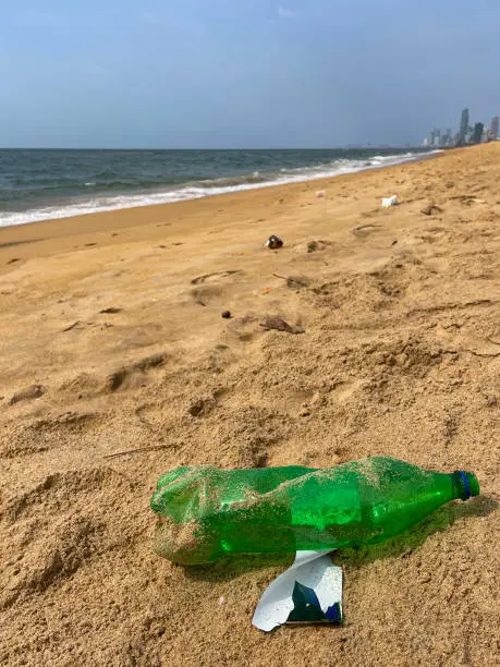 Photo of Close-up image of crushed, green plastic water bottle without lid washed up on sandy beach at low tide, ocean rubbish and pollution on shore, littering sand, dirty beach, coastline view of the skyscrapers of the city of Colombo, Mount Lavinia Beach