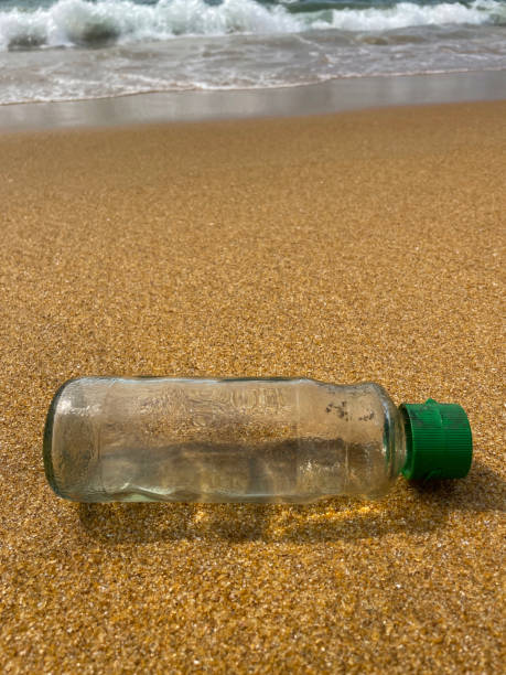 istock Close-up image of empty, transparent plastic drinks bottle with green lid washed up on sandy beach at low tide water's edge, breaking waves washing ocean rubbish and pollution on shore, littering sand, dirty beach, focus on foreground, copy space 1459142715