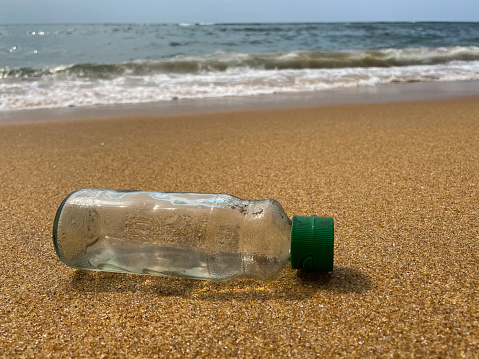 Stock photo of showing close-up view of sandy beach at low tide with empty, transparent plastic drinks bottle with green lid washed up from polluted sea.