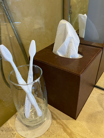 Stock photo showing close-up view of luxury bathroom marble counter with white toothbrushes in drinking glass on circular coaster and brown cube tissue box cover with white facial tissues in front of mirror.