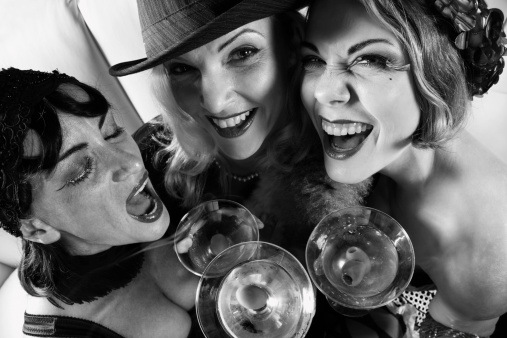 Retro style photograph of three young adult Caucasian females drinking martinis and smiling while looking up at viewer.