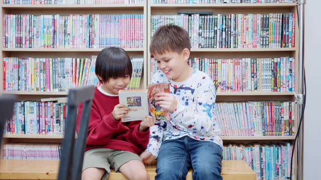 Two multiracial elementary schoolboys are sitting and talking about a comic book in the library at the school.