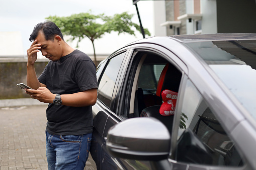 Sad and stressed asian online driver standing beside his car while holding a cell phone and his head