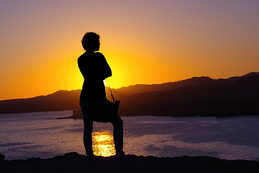 Silhouette of a woman traveler at sunset on the background of mountains and sea.