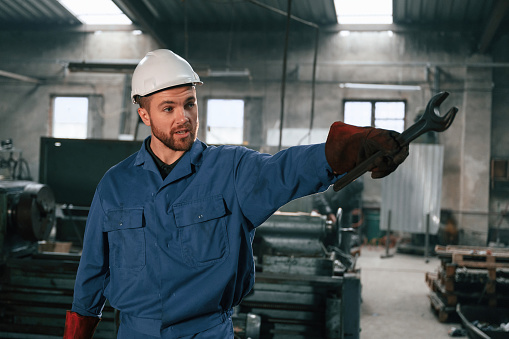 Metal wrench in hand. Factory worker in blue uniform is indoors.