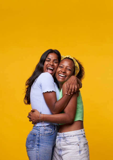 Vertical. Two diversity pretty positive girls toothy smile hug isolated on yellow color background. stock photo