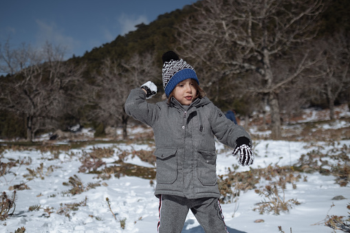 Portrait of adorable little kid boy with long blond hair playing with snowballs outdoors. Child with blue scarf and hat walking and having fun on a windy winter day.