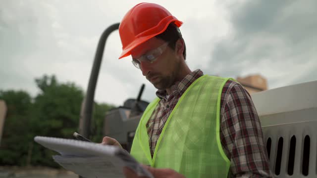 Engineer builder hard hat standing against paver at construction site, checking plan of project with technical documentation and using smartphone. Worker in front of roller on building site.