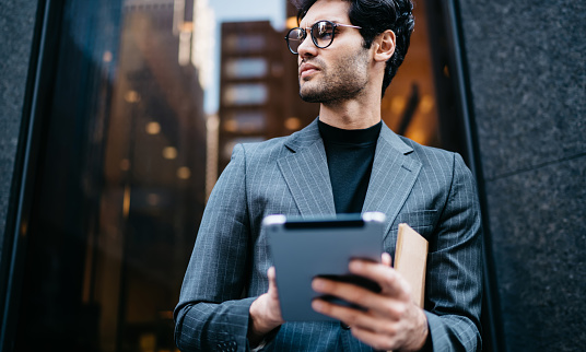 Businessman with bristles wearing eyeglasses standing in doorway of business center and holding tablet and copybook while pensively looking away