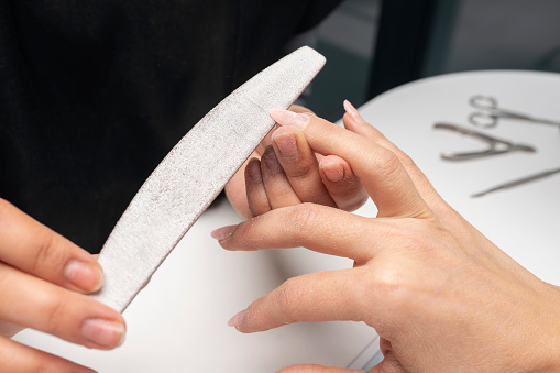 Manicurist is using a file in beauty salon. Cleaning and leveling the cuticle with nippers at the root of fingernails. Young woman getting a manicure. Beautiful hands and nails. Macro photo. Nail Care.