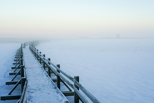 Hoarfrost and snow covered wooden boardwalk on a frozen lake, misty winter landscape
