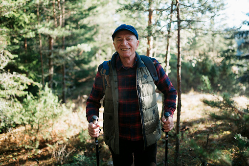 Smiling senior man walking on hiking trail through forest. Healthy mature man enjoying hiking in nature.