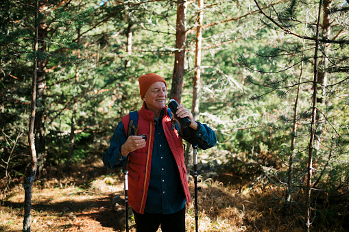 Active senior hiker drinking water from a bottle standing on forest trail with a backpack. Healthy man taking a break and drinking water while hiking in nature.