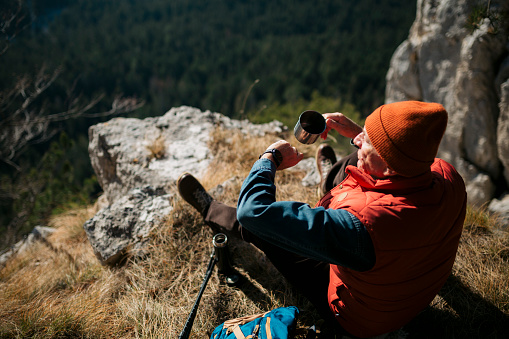 Senior man checking time on smartwatch while having coffee on mountain edge. Active senior male hiker taking a break while on hiking trip in nature.
