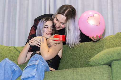 young long haired man hugging his partner from behind and giving her a gift for a special day at home. Celebration of Valentine's Day, birthdays, women's day...