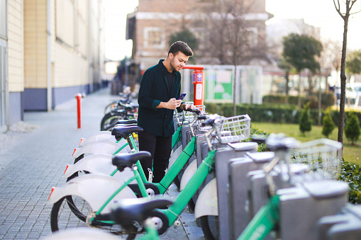 Young brown-haired man renting from bike rental station with his mobile phone. city ​​bike transportation concept. young man renting a bike in the city
