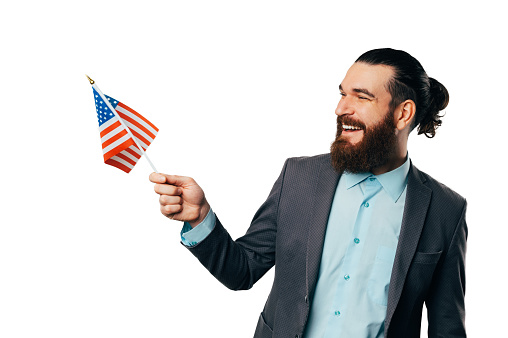 Young handsome cheerful bearded man wearing jacket and shirt holds the US flag over white background.