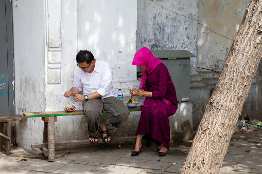 A couple eating street food on a wooden bench in the Kota Tua district of Jakarta, Indonesia.