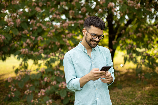 Young handsome male standing and smiling while sending message on his mobile phone outdoor