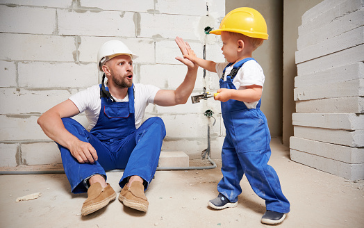 Male builder and little boy slapping hands giving high five in room with brick wall. Kid in safety helmet holding wire stripper cutter tool and celebrating successful home renovation with father.