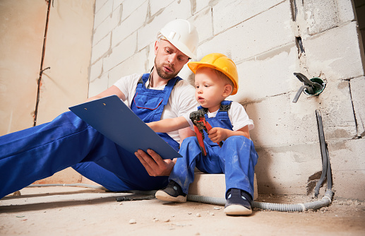 Male worker sitting by brick wall and pointing at clipboard while little boy holding tool. Man and kid wearing safety helmets and work overalls while working with documents at construction site.