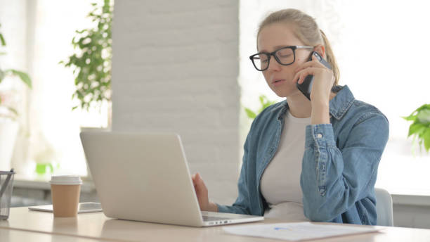 istock Beautiful Young Woman Talking on Phone while using Laptop in Office 1459197518