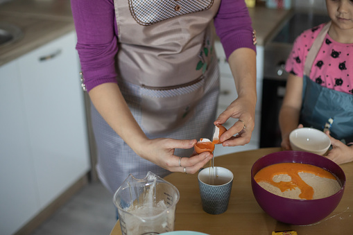 Close up of young woman and her daughter while they making a cake in the kitchen. Young woman breaks an egg to make a dough.