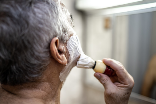 Close-up of a senior man passing shaving foam on face in front of mirror in bathroom at home