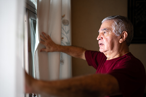 Worried senior man looking through window at home