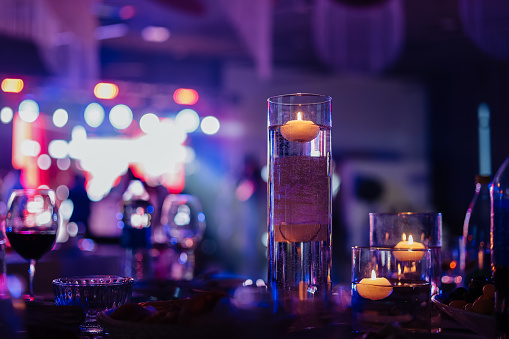 Banquet table decorated with burning candles in glass vases in restaurant hall. In the background party with silhouettes of people dancing on the dance floor with disco lights glowing searchlight.
