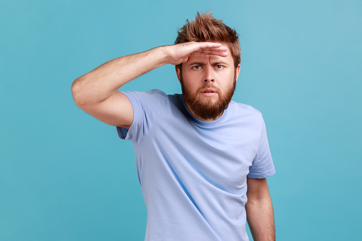 Portrait of serious concentrated bearded man looking far away at distance with hand over head, attentively searching for bright future. Indoor studio shot isolated on blue background.