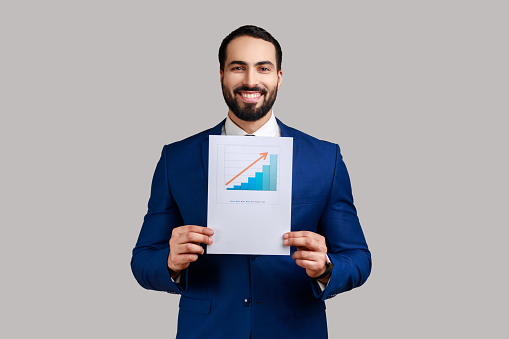 Handsome bearded businessman standing showing business growth graph, looking at camera with happy expression, wearing official style suit. Indoor studio shot isolated on gray background.