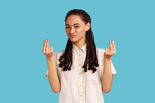 Portrait of beautiful woman with black dreadlocks makes money gesture, rubs fingers, looking at camera with glad expression, wearing white shirt. Indoor studio shot isolated on blue background.