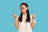 Woman with dreadlocks makes money gesture, rubs fingers, looking at camera with glad expression.