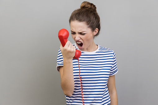 Portrait of angry nervous woman wearing striped T-shirt screaming and yelling talking retro landline phone, complaining on connection quality. Indoor studio shot isolated on gray background.