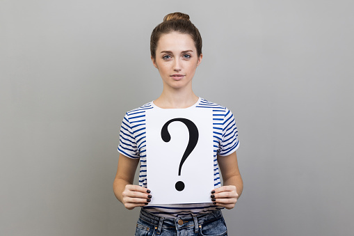 Portrait of serious attractive dark haired woman wearing striped T-shirt holding paper with question mark, thinks about tasks. Indoor studio shot isolated on gray background.