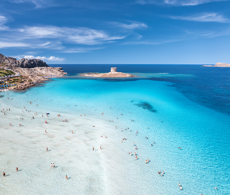 Aerial view of famous La Pelosa beach at sunny summer day. Stintino, Sardinia island, Italy. Top view of sandy beach, swimming people, clear blue sea, old tower and sky with clouds. Tropical seascape