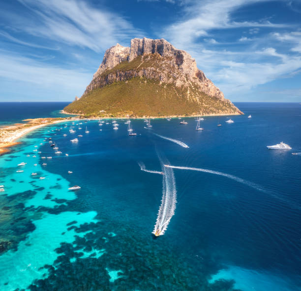 Aerial view of luxury yachts and boats on blue sea, beautiful mountain and sky with clouds at summer sunny day. Sandy beach on Tavolara island in Sardinia, Italy. Top view. Colorful seascape. Travel stock photo