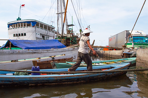 Jakarta, Indonesia - November 6, 2010.\nA man on a small wooden boat beside Pinisi ships at the Port of Sunda Kelapa in Jakarta, Java, Indonesia on the Ciliwung river. These days the port only accommodates old wooden trading boats.