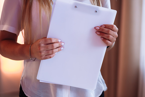cropped photo of young woman holding a magazine or folder with mock up. girl in a white shirt standing near the window indoors