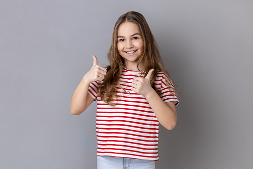 Little girl wearing striped T-shirt looking at camera with toothy smile and showing thumbs up, approval sign, satisfied with service, good feedback. Indoor studio shot isolated on gray background.