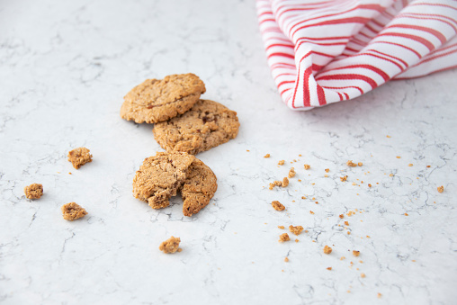 Several oatmeal cookies on kitchen counter with different sizes of crumbs. Kitchen counter with white and red striped kitchen towel.