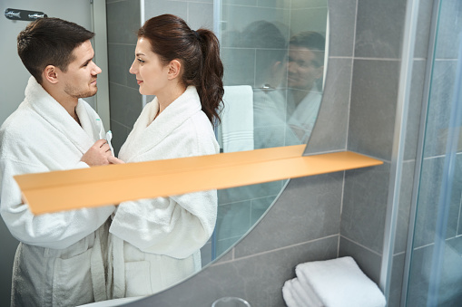 Man and a woman in bathrobes stand in bathroom and look at each other, they have toothbrushes in their hands