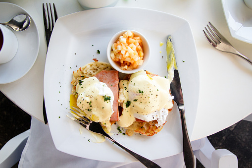 A white plate of Eggs Benedict and a cup of diced cantaloupe fruit for breakfast or brunch. Set in a restaurant with a white table and silverware.